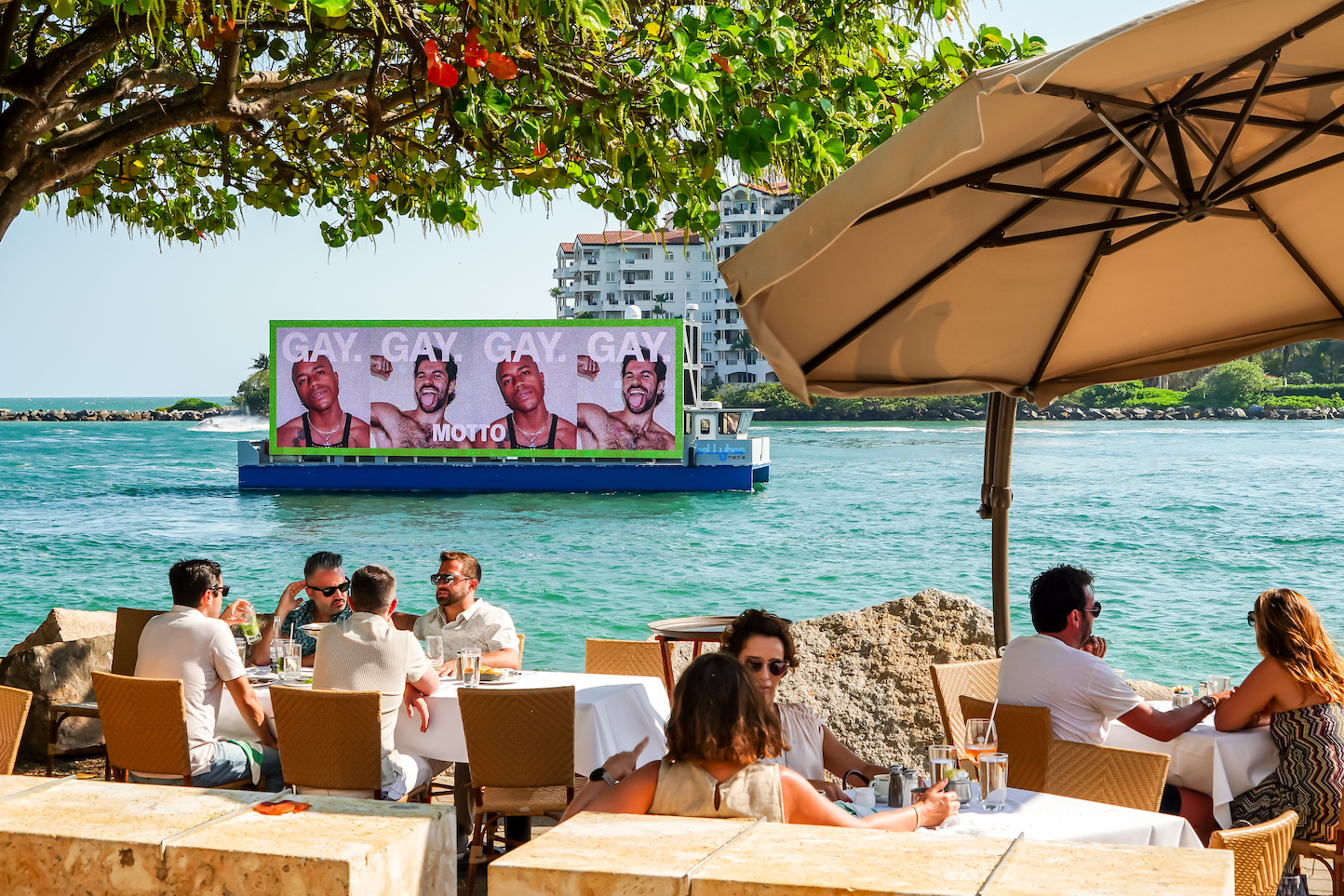 A boat with a digital billboard of the Motto campaign seen in the distance behind a group of people at an outdoor cafe